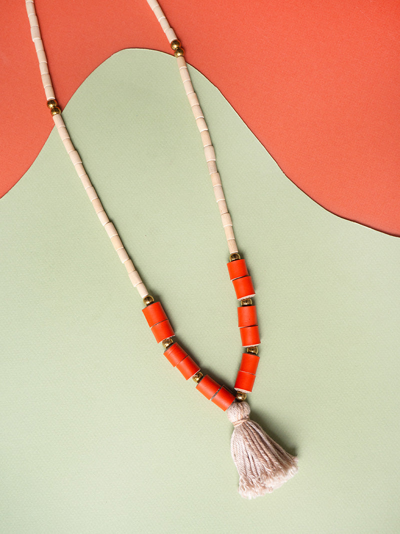 Collar largo de piezas de madera natural con rollitos en cuero color naranja, bolitas doradas de acero inoxidable y rollito de hilos de algodón en gris teñidos con tintes naturales de frijoles negros.