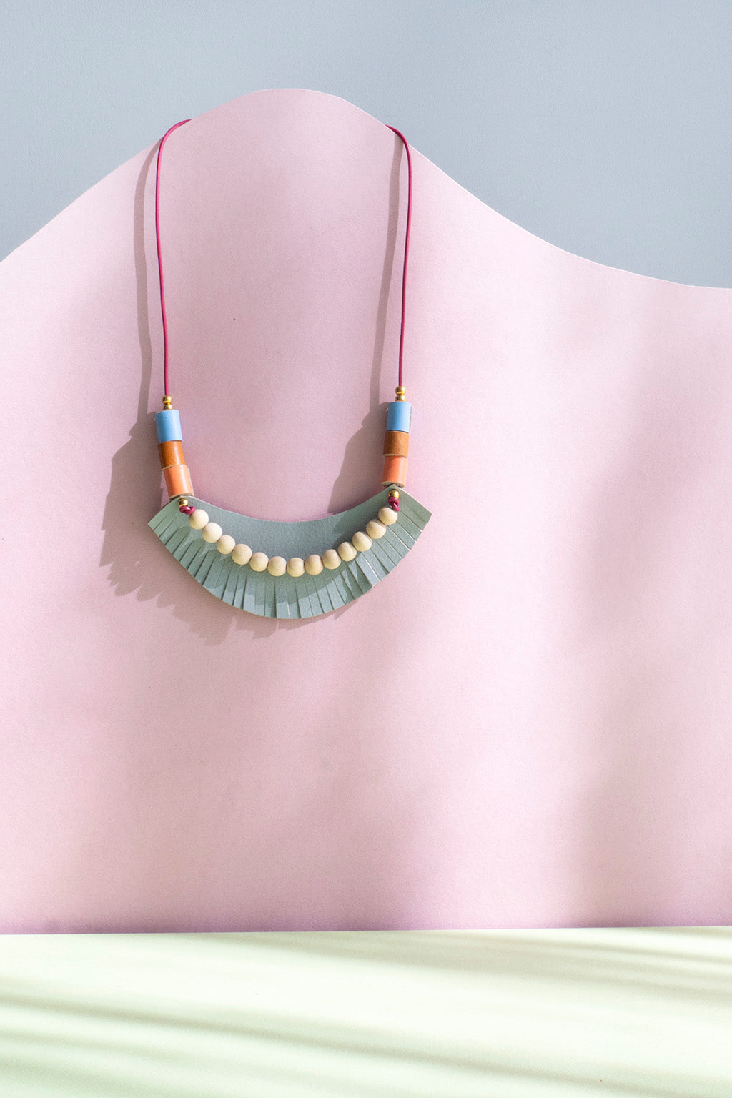 Collar ajustable, pieza de cuero gris con barbas, sobre ella bolitas de madera natural junto a rollitos en cuero durazno, miel y cielo, con bolitas doradas de acero inoxidable, sobre fondo de color rosado y verde claro.