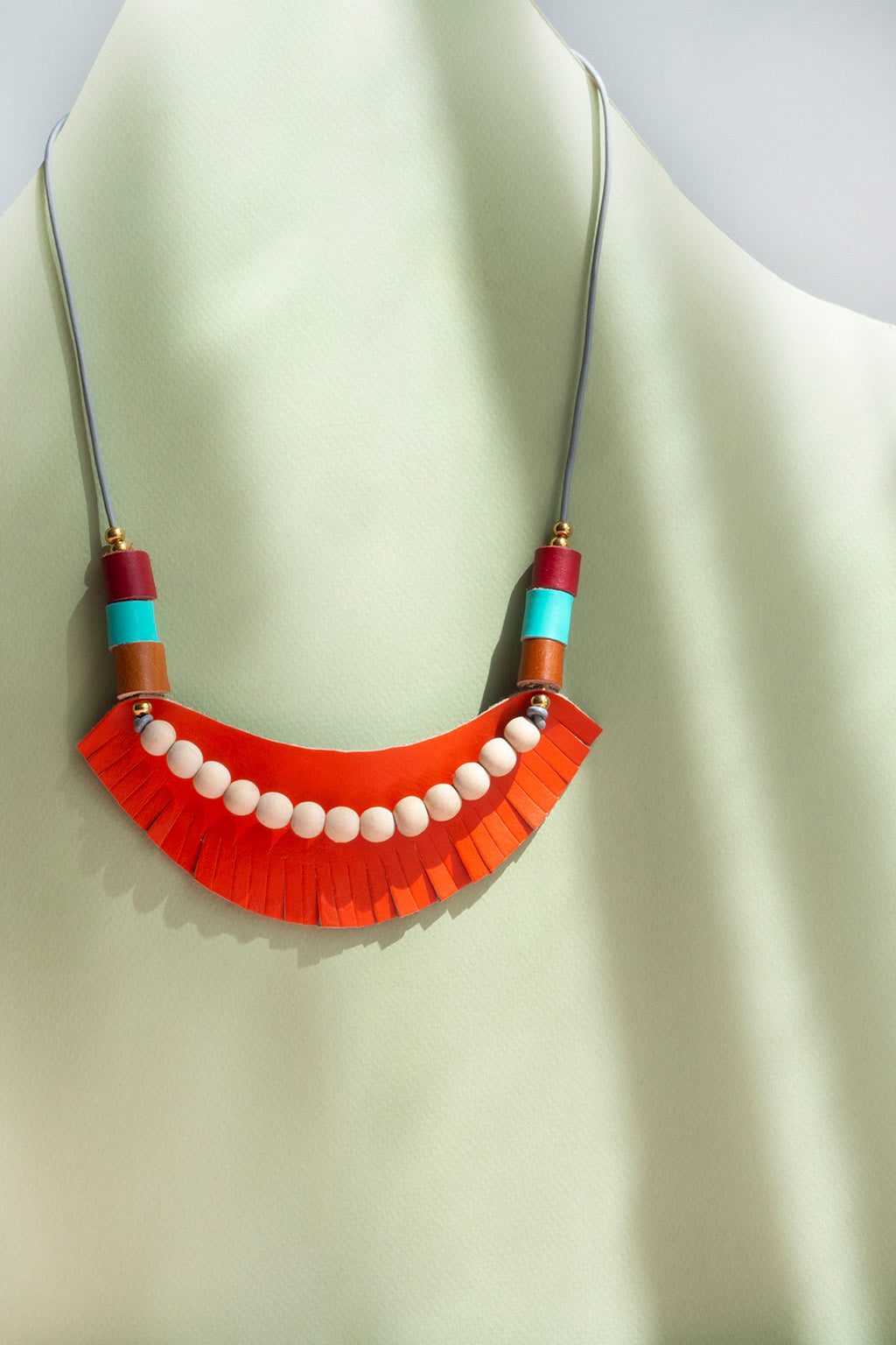 Collar ajustable, pieza de cuero naranja con barbas, sobre ella bolitas de madera natural junto a rollitos en cuero miel, turquesa y rojo, con bolitas doradas de acero inoxidable, sobre fondo de color verde claro.
