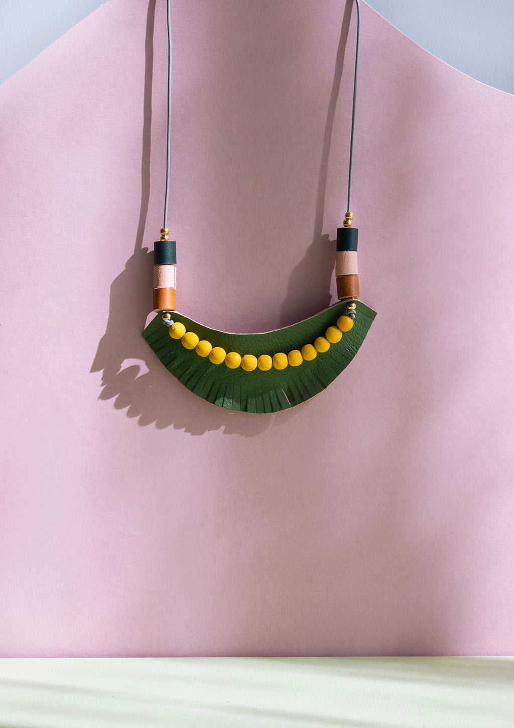 Collar ajustable, pieza de cuero verde olivo con barbas, sobre ella bolitas de madera natural en color amarillo junto a rollitos en cuero miel, salmón y verde inglés, con bolitas doradas de acero inoxidable, sobre fondo de color rosado y verde claro.