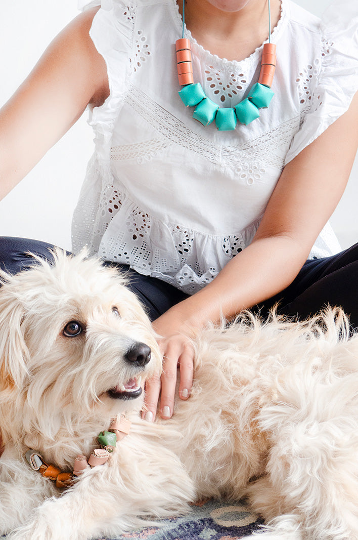 Mujer sentada con blusa blanca y collar cubos en colores turquesa y durazno acompañada de perrita blanca con ojos cafés
