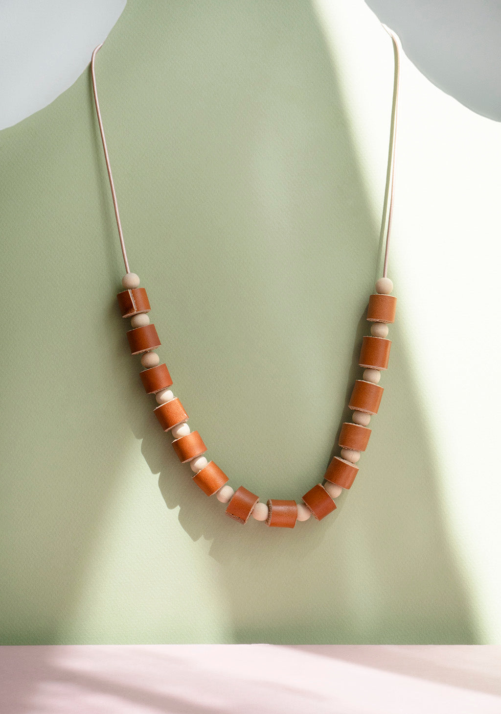 Collar rollitos miel con bolitas de madera natural con cordón de cuero rose gold que guinda sobre un fondo de color verde oliva y rosado.