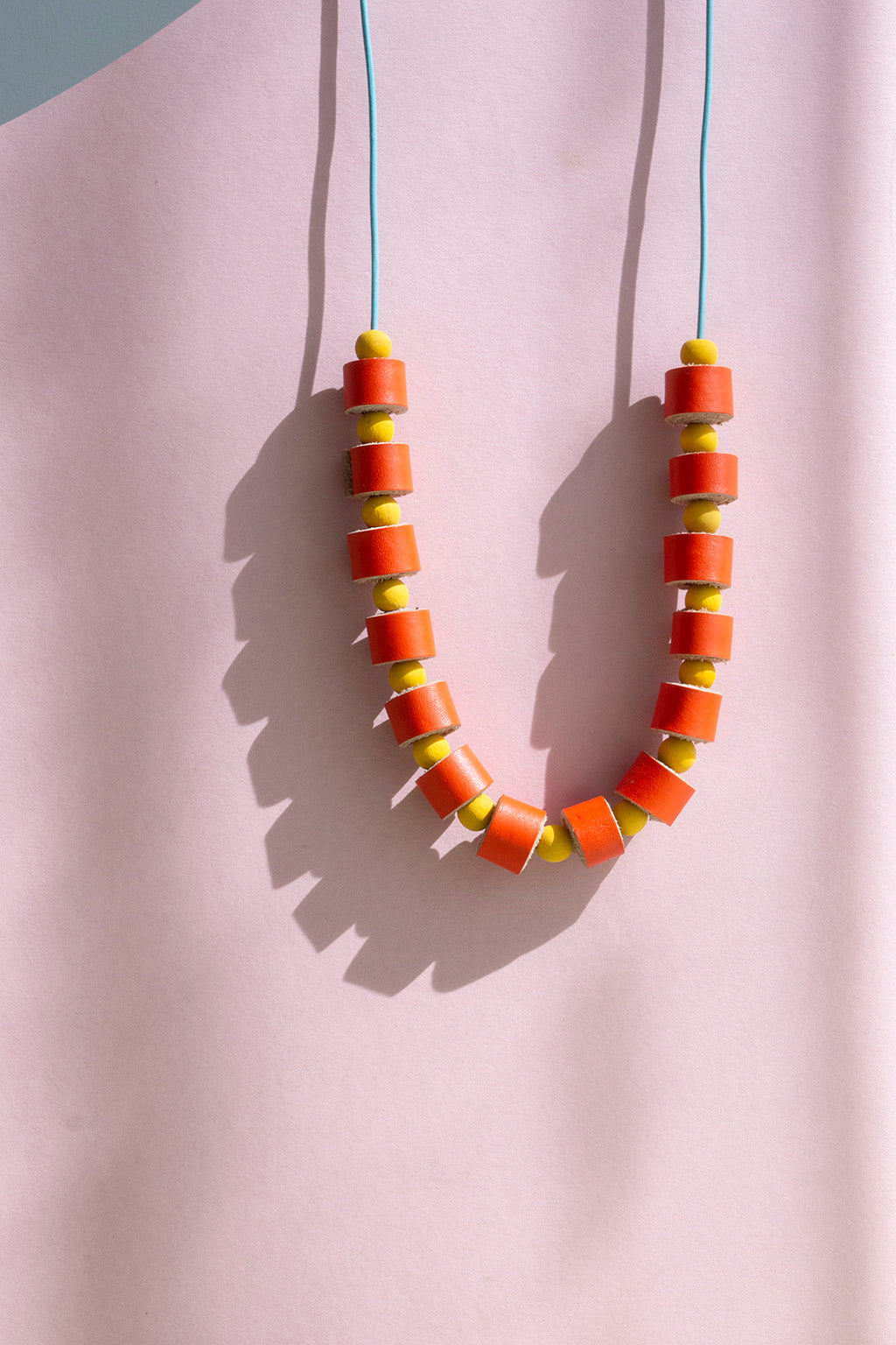 Collar rollitos naranja con bolitas de madera amarillas con cordón de cuero celeste que guinda sobre un fondo de color rosado