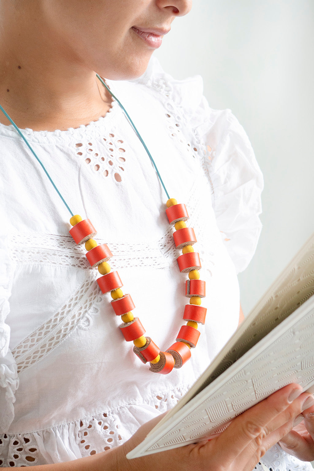 Mujer con blusa blanca usando el collar helena en colores naranja y amarillo. La mujer sostiene un libro abierto en sus manos.
