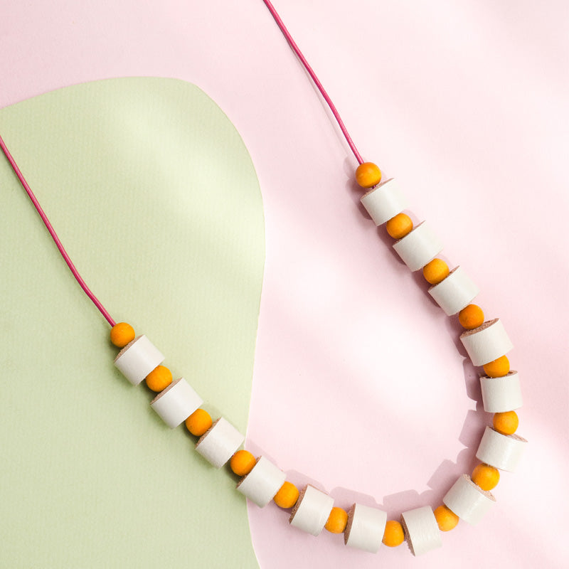 Collar rollitos vainilla con bolitas de madera amarillas con cordón de cuero vino que guinda sobre un fondo de color rosado y verde oliva.