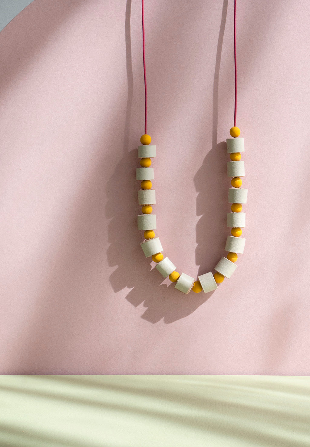 Collar rollitos vainilla con bolitas de madera amarillas con cordón de cuero vino que guinda sobre un fondo de color rosado y verde oliva.