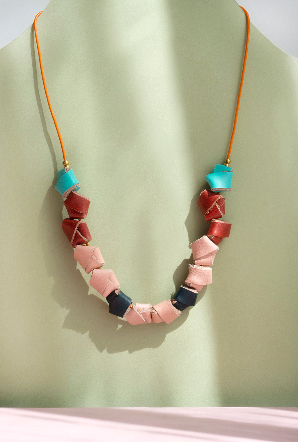 Collar de colores turquesa, rojo, salmón y verde inglés con bolitas doradas y cordón naranja guindando sobre fondo verde claro y rosado